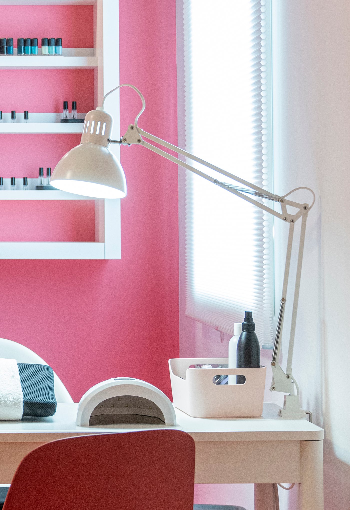Skrivbord med vit TERTIAL arbetslampa, förvaringslåda och flaskor mot en rosa vägg. Hylla med nagellack i bakgrunden och fönster med persienner ger ett ljust arbetsutrymme.

