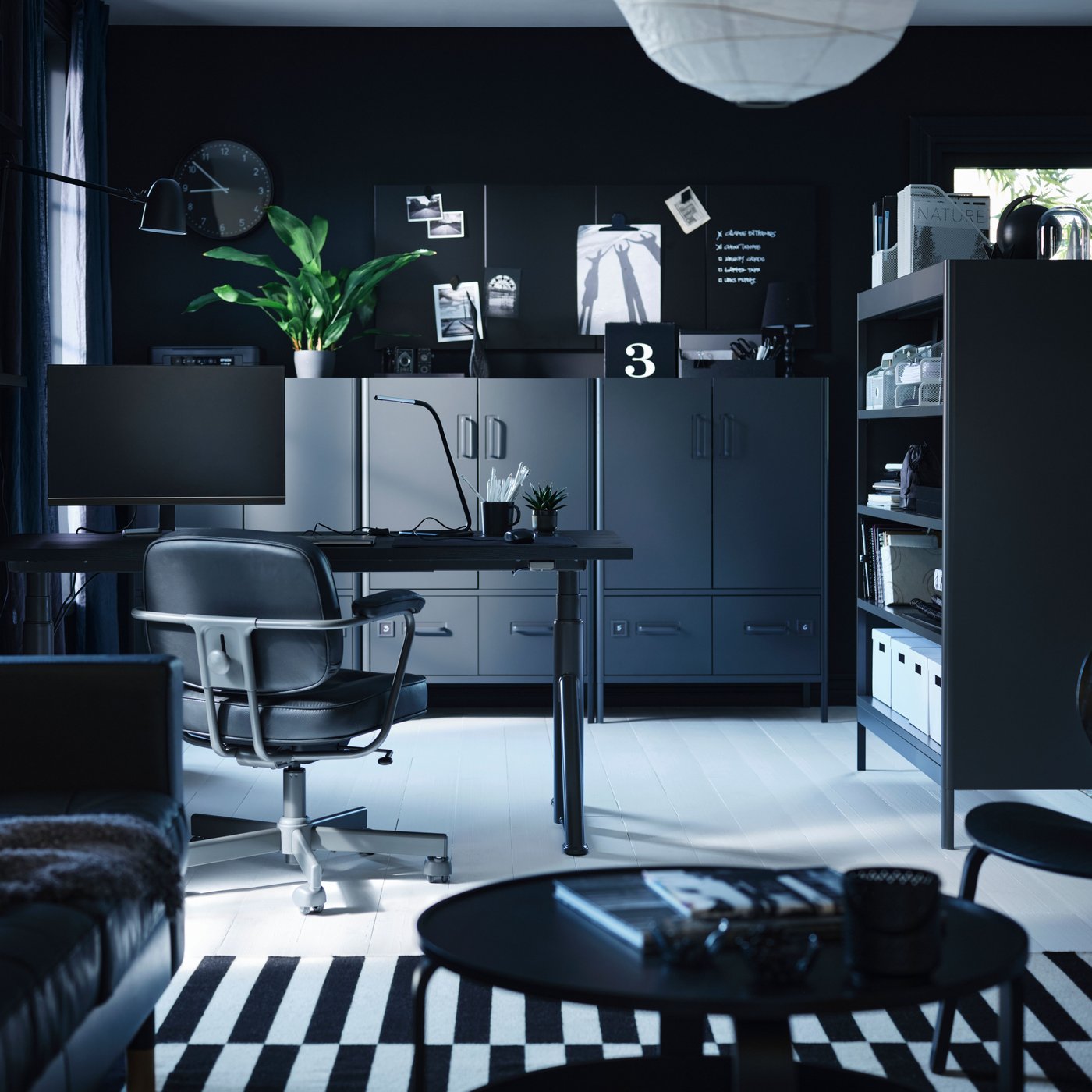 Een stijlvolle IKEA thuiswerkplek beschikt over een zwarte JÄRVFJÄLLET bureaustoel, een zwart BEKANT zit-sta bureau met een grote monitor en een FORSÅ bureaulamp, alles geplaatst tegen donkere muren met een plant en een wandklok.