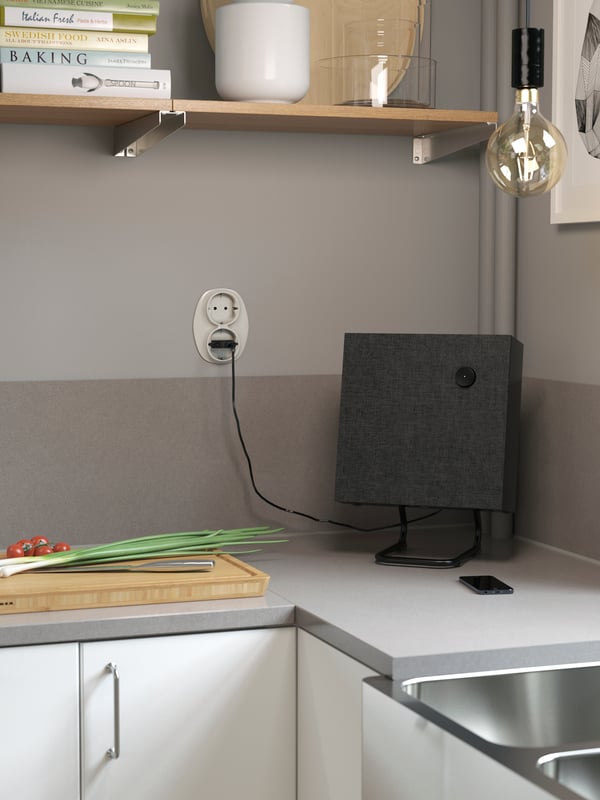A square speaker standing on a grey worktop with matching splashback. A wall-mounted shelf and cord set with bulb are above.