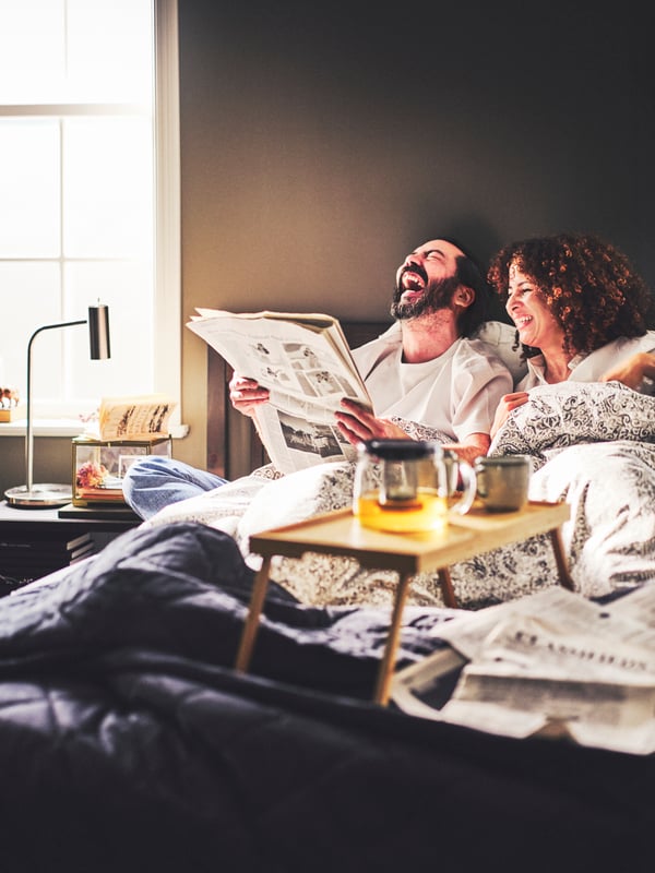 Two people in a bed dressed in a ÄNGSKLOCKA duvet cover with 2 pillowcases with a RESGODS bed tray, and a lamp to the side.