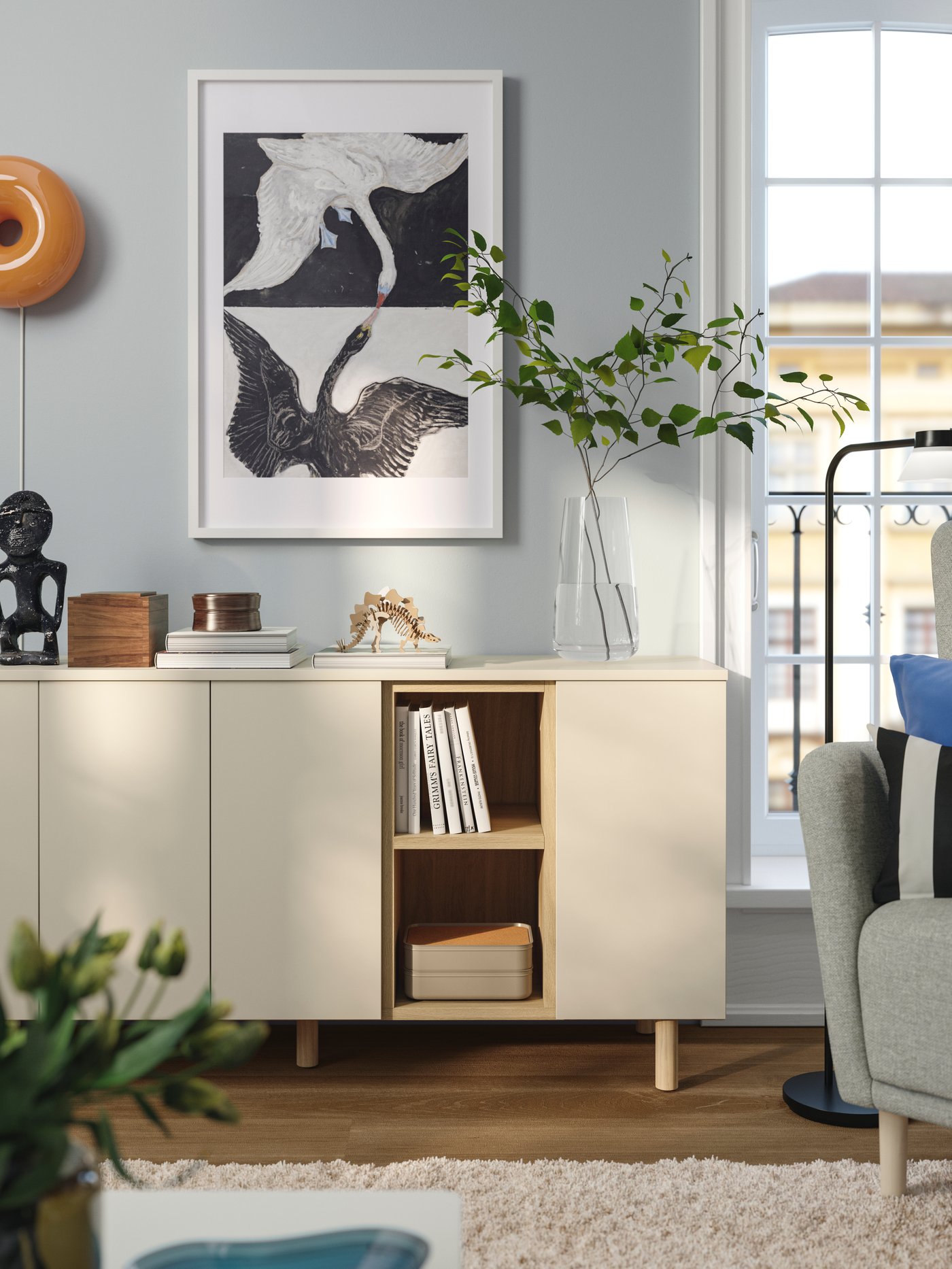 A clutter-free reading corner with light grey-beige LEDAMOT cabinets, a grey armchair, books, art and fresh greenery.