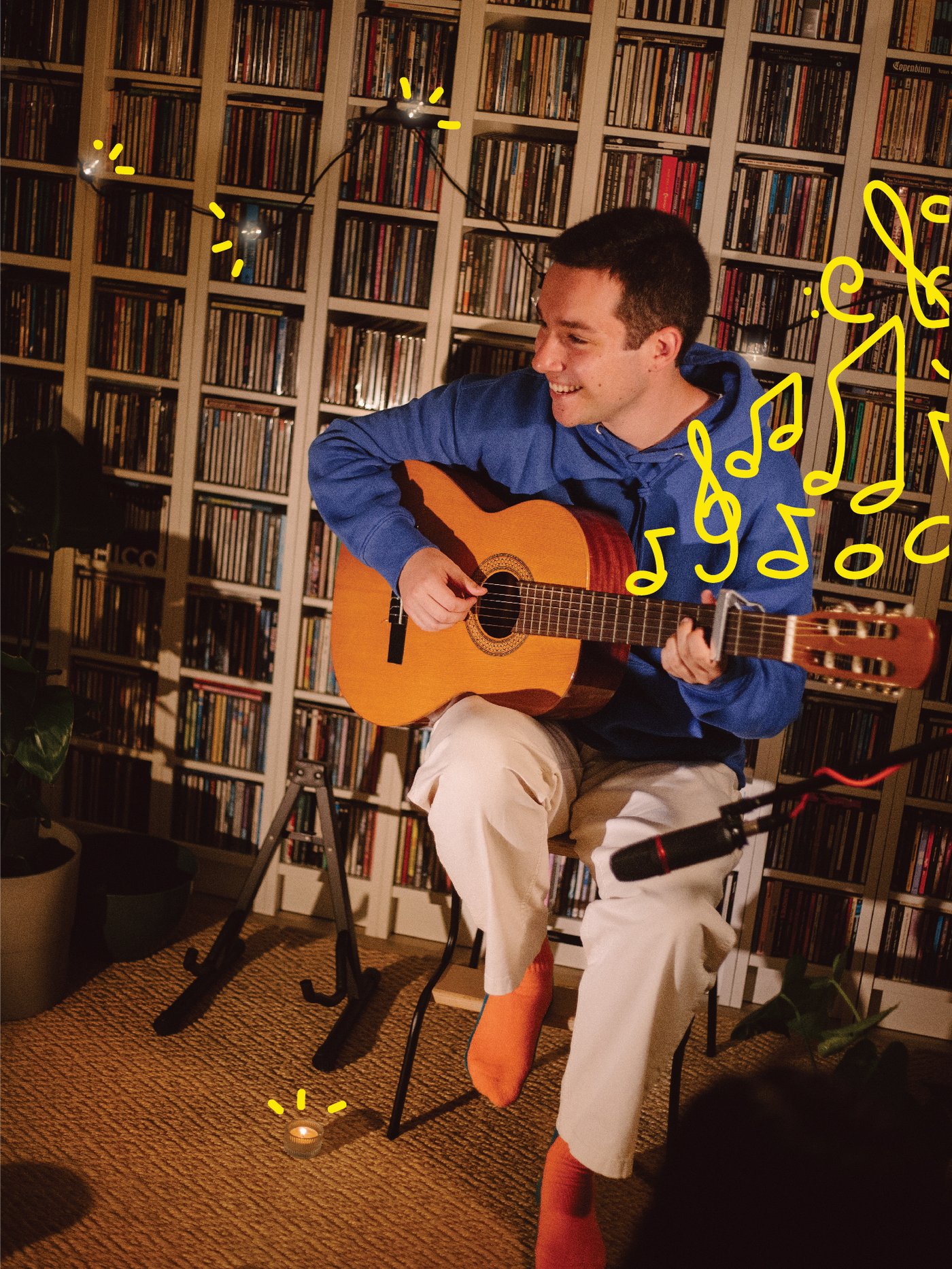 A man playing the guitar in front of a bookcase wall decorated with string lights.