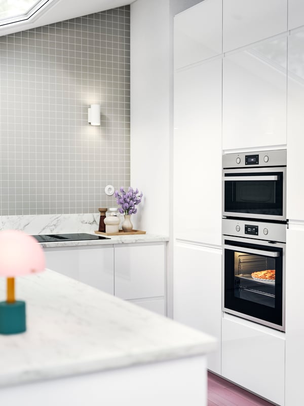 VOXTORP kitchen with white high-gloss fronts, a pizza in the oven, and a tiled backsplash with a soft grey tone.