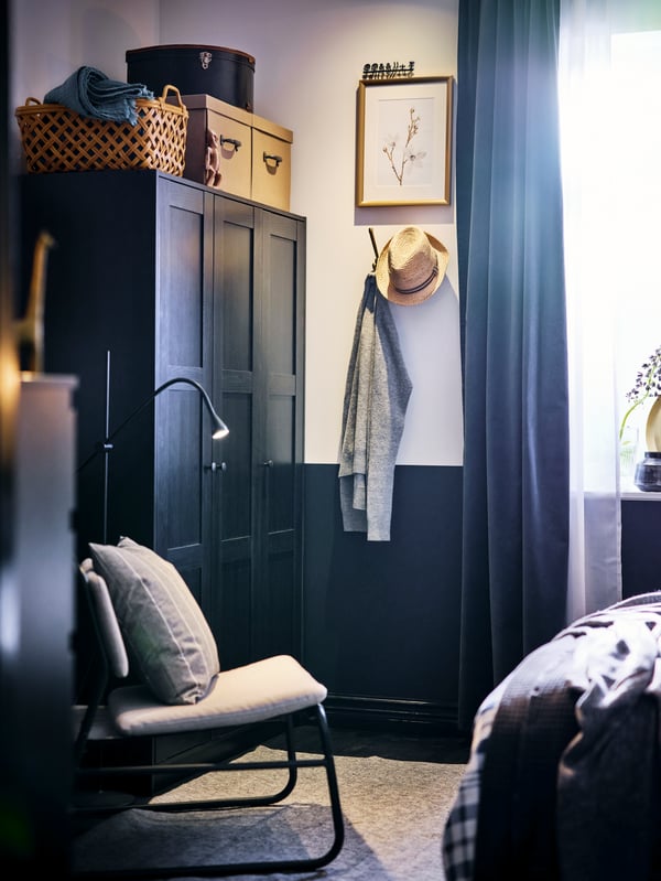 Le coin d’une chambre à coucher avec une armoire-penderie RAKKESTAD noir-brun, un lampadaire et un fauteuil relax LINNEBÄCK gris pâle.
