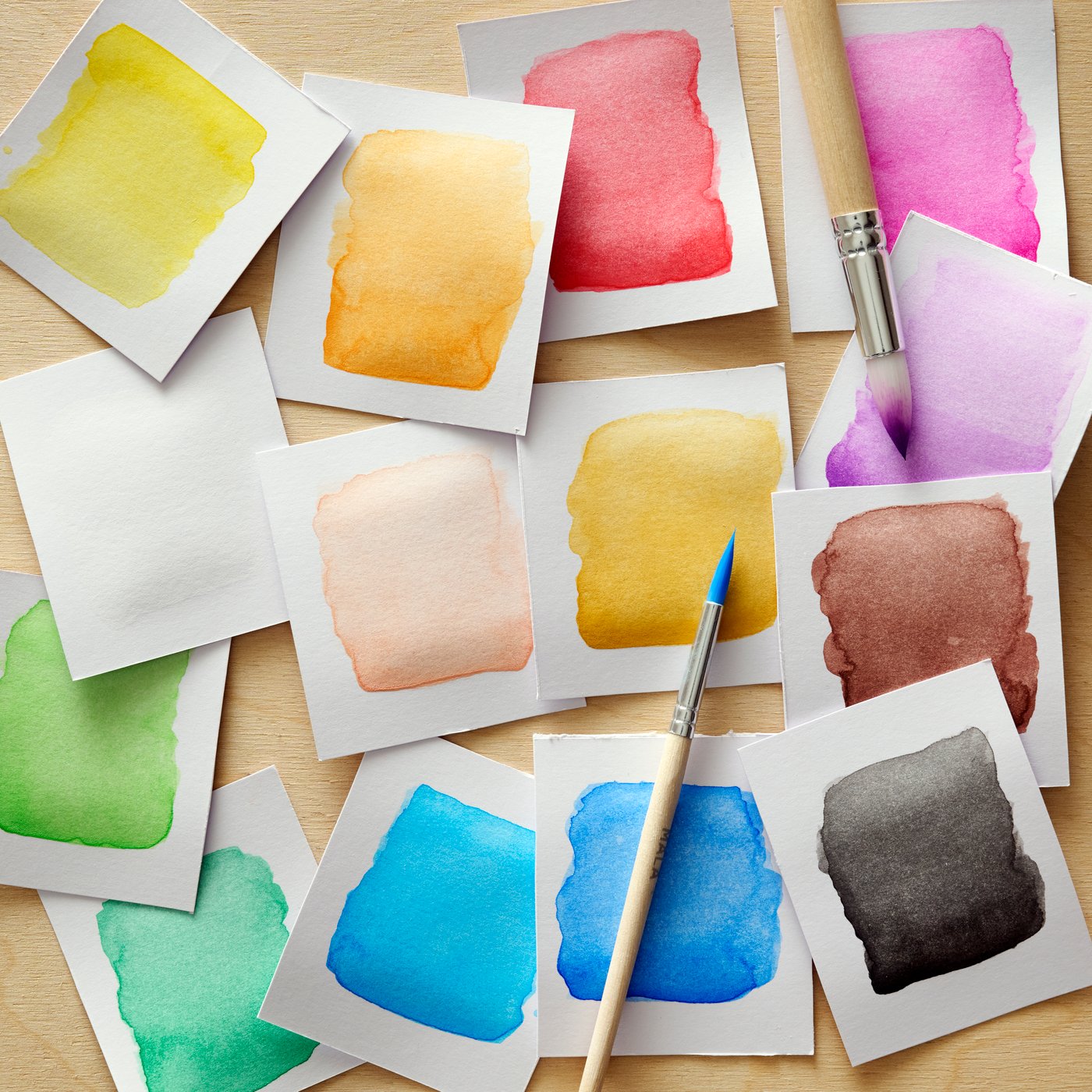 Watercolor paint swatches in various colors scattered on a surface with two paintbrushes placed among them.