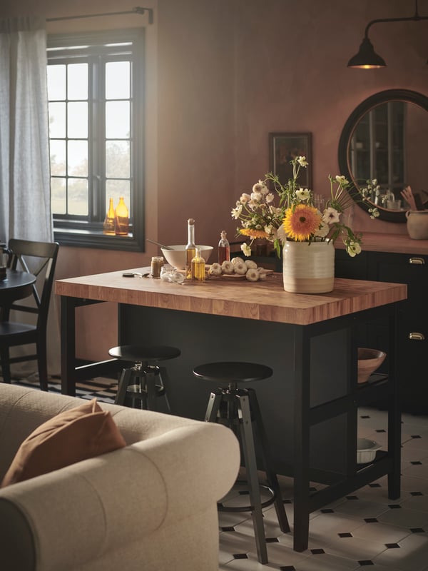 Several DALFRED bar stools placed around a VADHOLMA kitchen island with black legs and a dark wood accent top. A VISKAFORS beige sofa sits infront of the kitchen island.