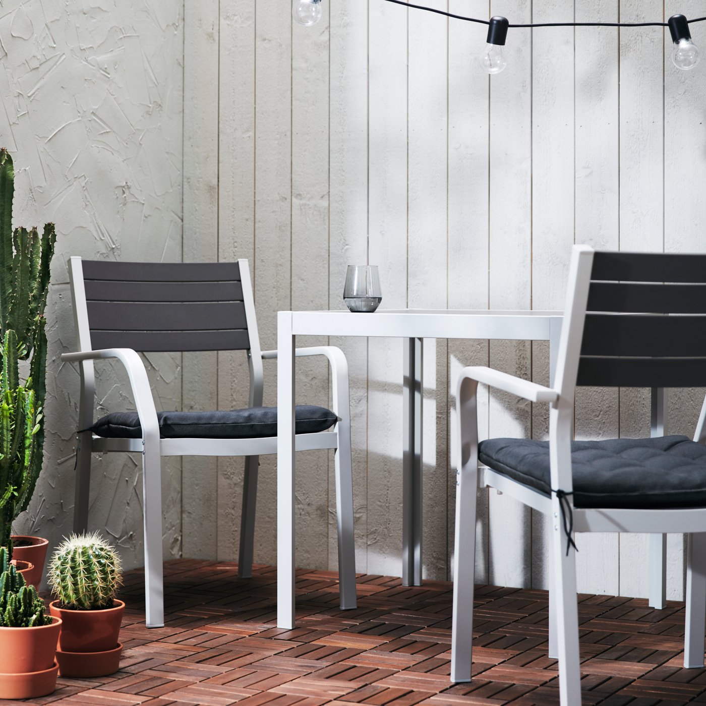 An outdoor table in white, with two chairs in white with dark grey upholstery and potted plants of different types.