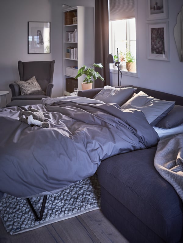 A dark grey GRÖNLID 3-seat sofa bed with chaise longue made up as a bed with grey bedding in a room with a half-open blind.