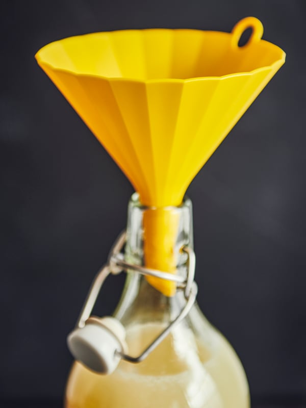 An UPPFYLLD funnel in bright yellow placed in the neck of a KORKEN bottle with stopper in clear glass, liquid in the bottle.