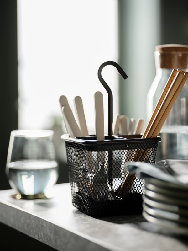 An anthracite-coloured UPPDATERA cutlery caddy, cutlery and TREBENT chopsticks, next to a glass carafe and a stack of plates.