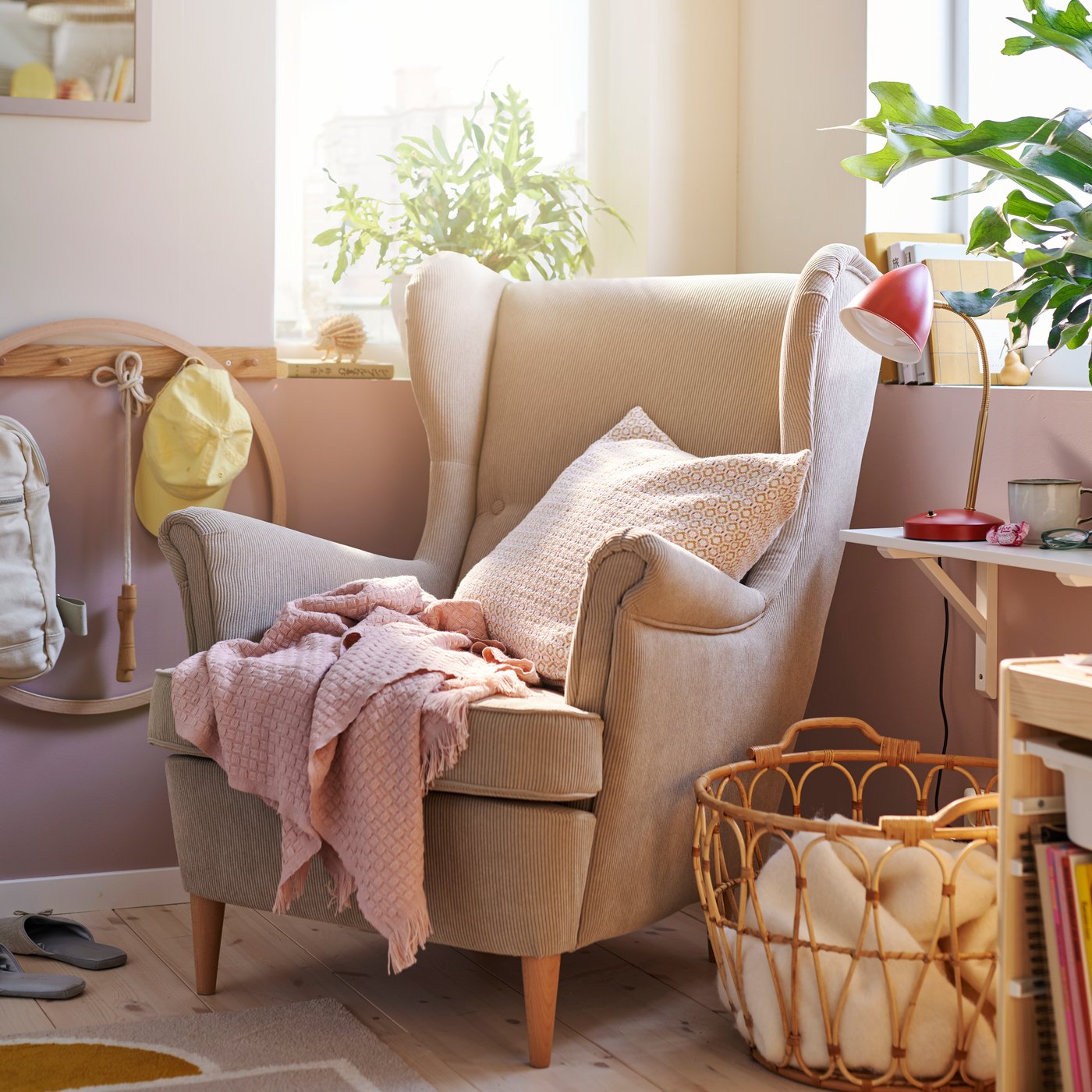 A pink HORNMAL throw beside a cushion in a light pink MOROTSMOTT cover on a beige STRANDMON wing chair in a sunny corner.
