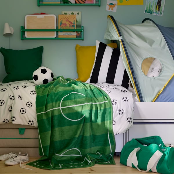 Ein grünes SPORTSLIG-Badetuch mit weißem Fußballfeldmuster liegt über einem Bett in einem Kinderzimmer.