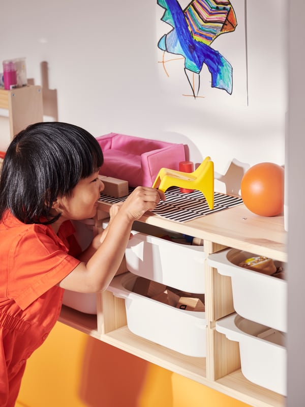A child picks a toy off the top of a light white stained pine TROFAST wall storage filled with white storage boxes.