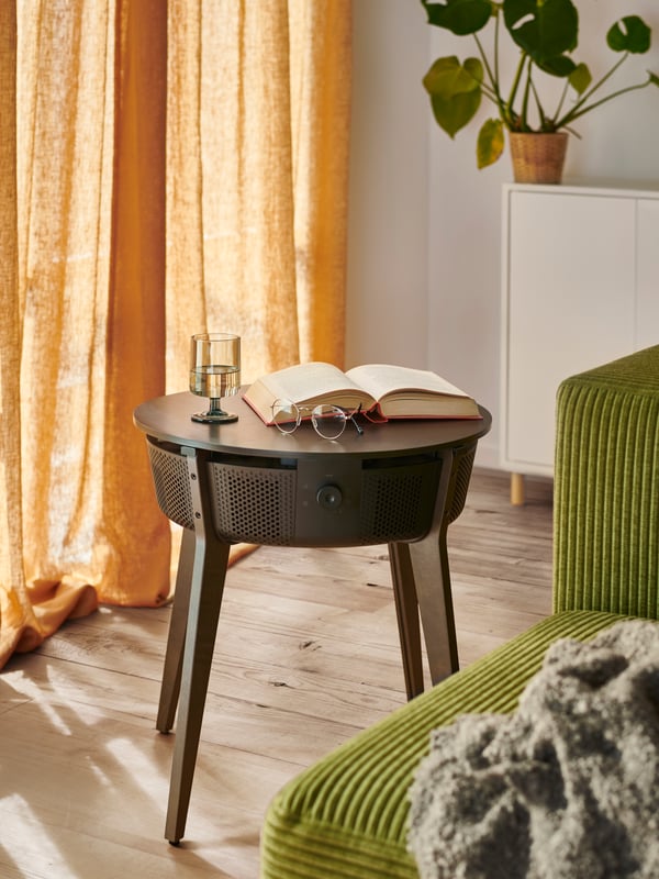 Une table STARKVIND avec un purificateur d’air est dans une pièce au sol en bois baigné par une lumière naturelle tamisée qui passe à travers des rideaux DYTÅG beige foncé.