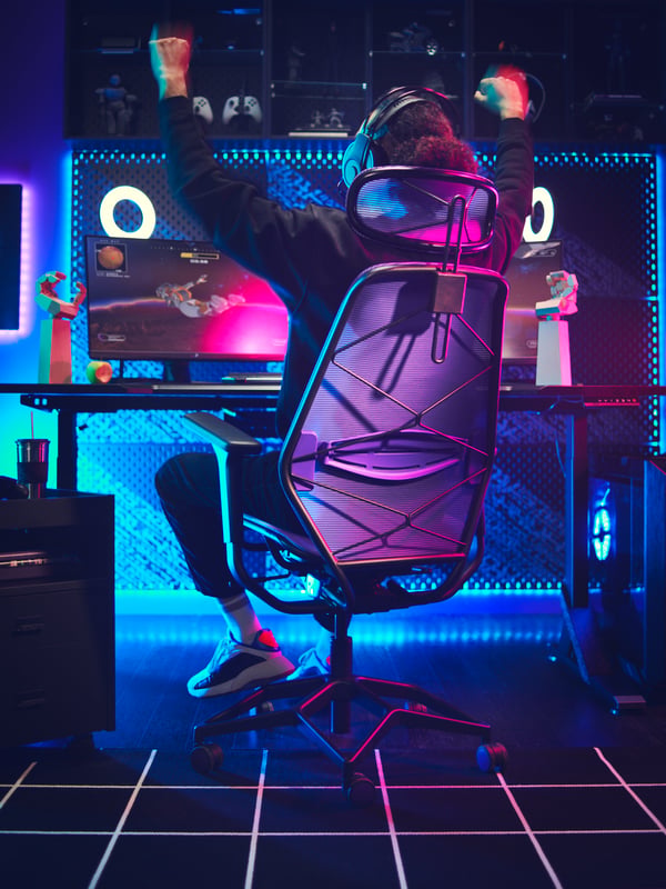 Une pièce sombre éclairée au néon avec une personne portant un casque assise dans une chaise de jeu STYRSPEL pourpre et noire, les bras levés pour célébrer une victoire.
