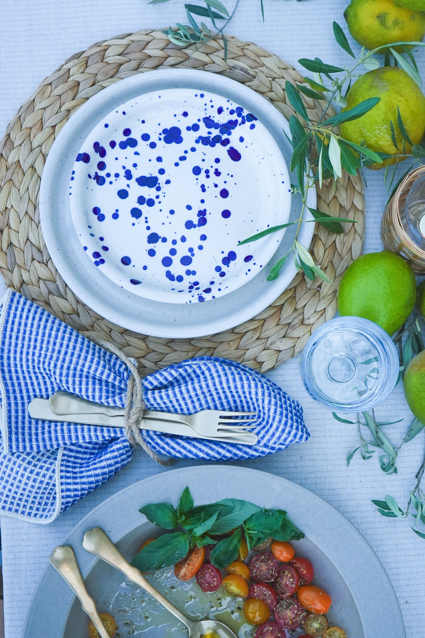 Mediterrane Tischdekoration mit blau-weissem SILVERSIDA Geschirr auf Bast-Platzset, Besteck in karierter Serviette, frische Limetten und Kirschtomaten-Basilikum-Salat.