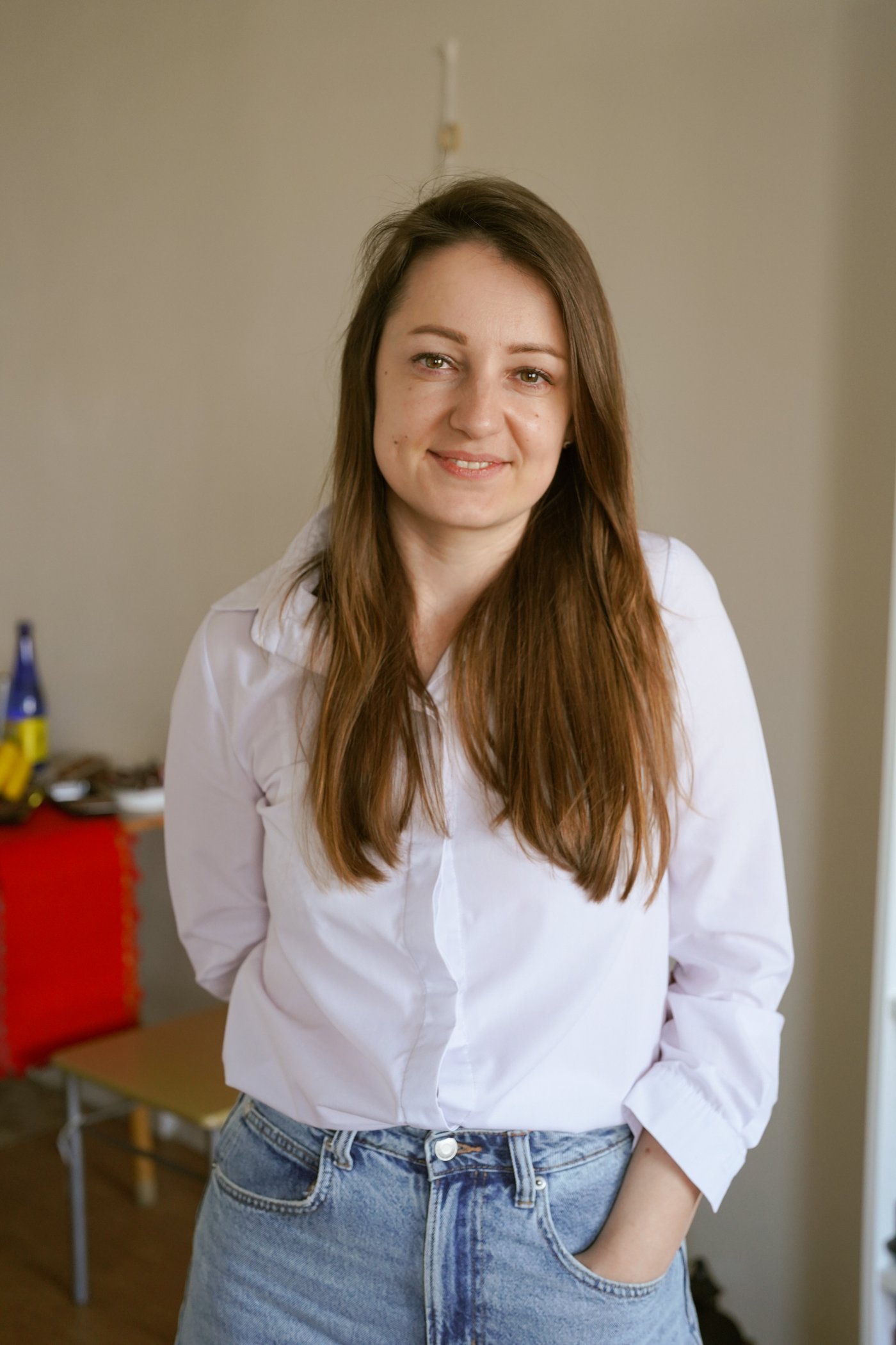 Devojka stoji unutra i gleda u kameru u beloj košulji i farmerkama, sa smeđom puštenom kosom i rukom u džepu.