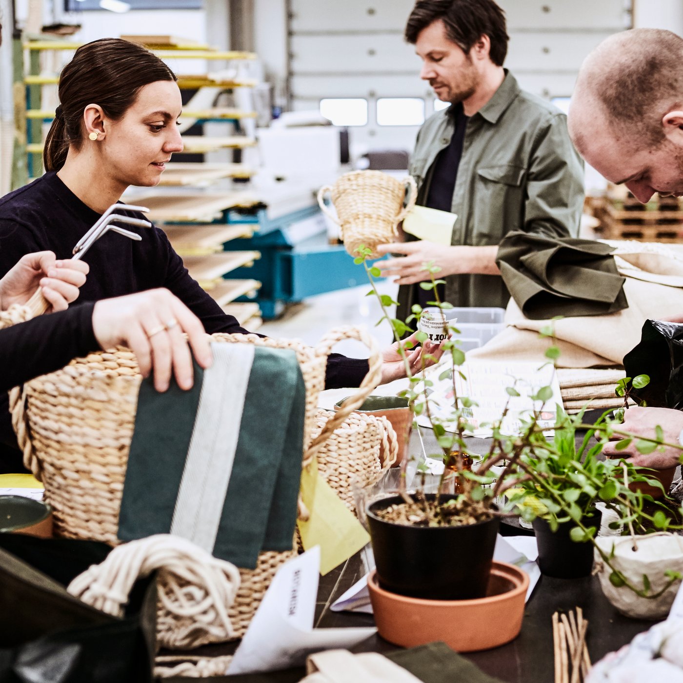 Fire IKEA designere inspicerer vævede kurve omkring en stor bordplade fyldt med foldet materiale og potteplanter.