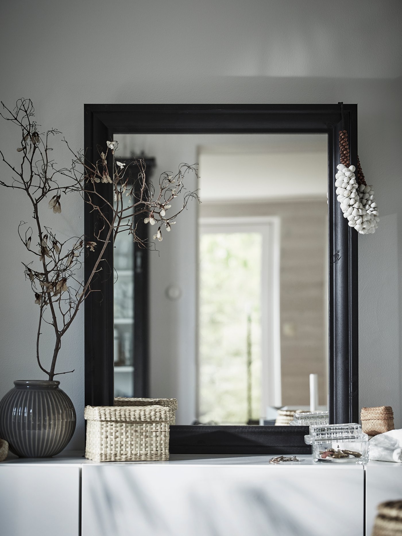 Miroir TOFTBYN noir, vase avec une branchette séchée, petite boîte de rangement et accessoires sur un buffet blanc.