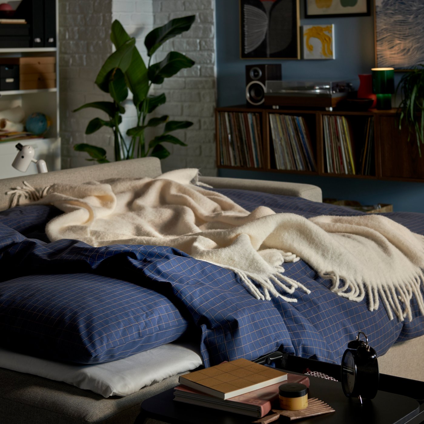 A cosy bed with blue-checked bedding, a white throw, and a green, leafy potted plant in a bright bedroom setting.