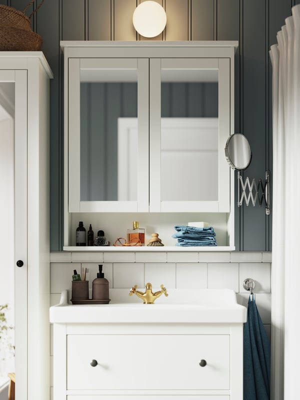 A FRIHULT wall lamp in metal and glass is mounted above a white HEMNES mirror cabinet and a white HEMNES wash-stand.