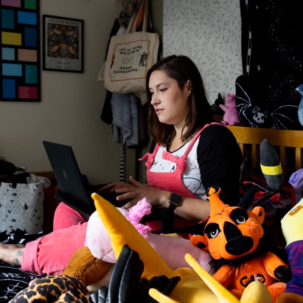 Lady sitting on her bed surrounded by plush toys, looking at her laptop.