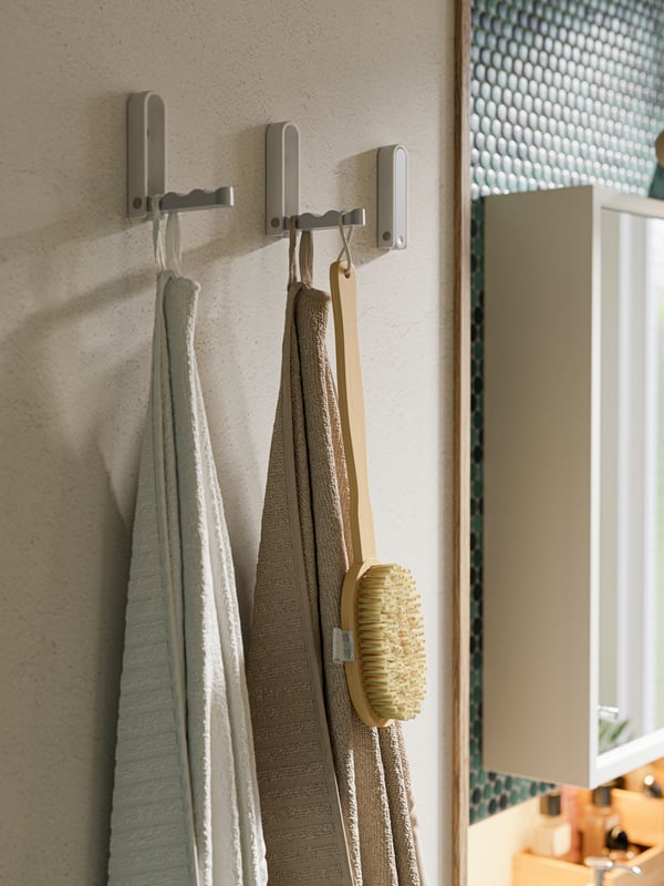 Three folding hooks on a wall, a light beige bath towel, a white bath towel and a wooden shower brush.