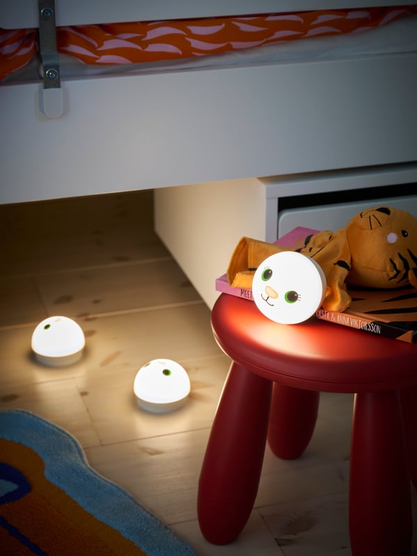 Gros plan de la zone près et sous le lit, avec trois veilleuses LED KORNSNÖ sous le lit et sur un tabouret rouge MAMMUT.