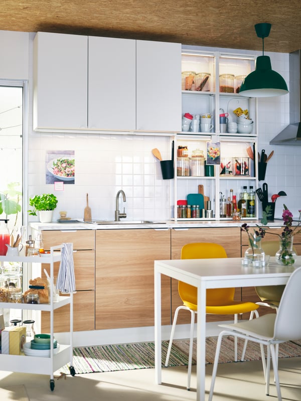 Una cocina con gabinetes ENHET, una mesa blanca MELLTORP, sillas LEIFARNE de diferentes colores y un carrito blanco NISSAFORS.