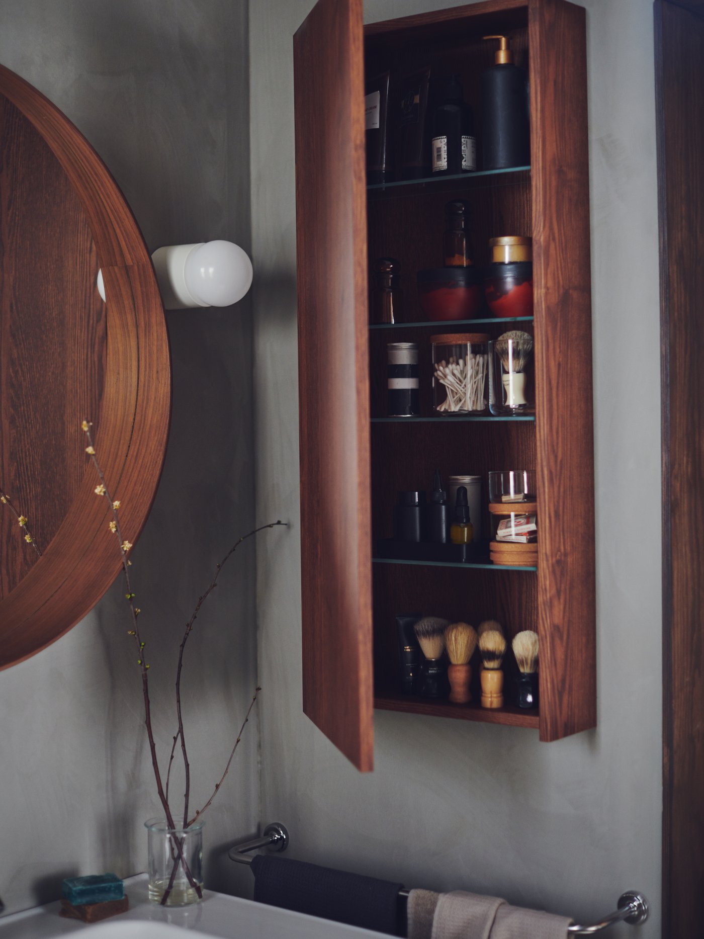 A brown GODMORGON cabinet on a bathroom wall with an open door showing shelves holding toiletries and shaving accessories.