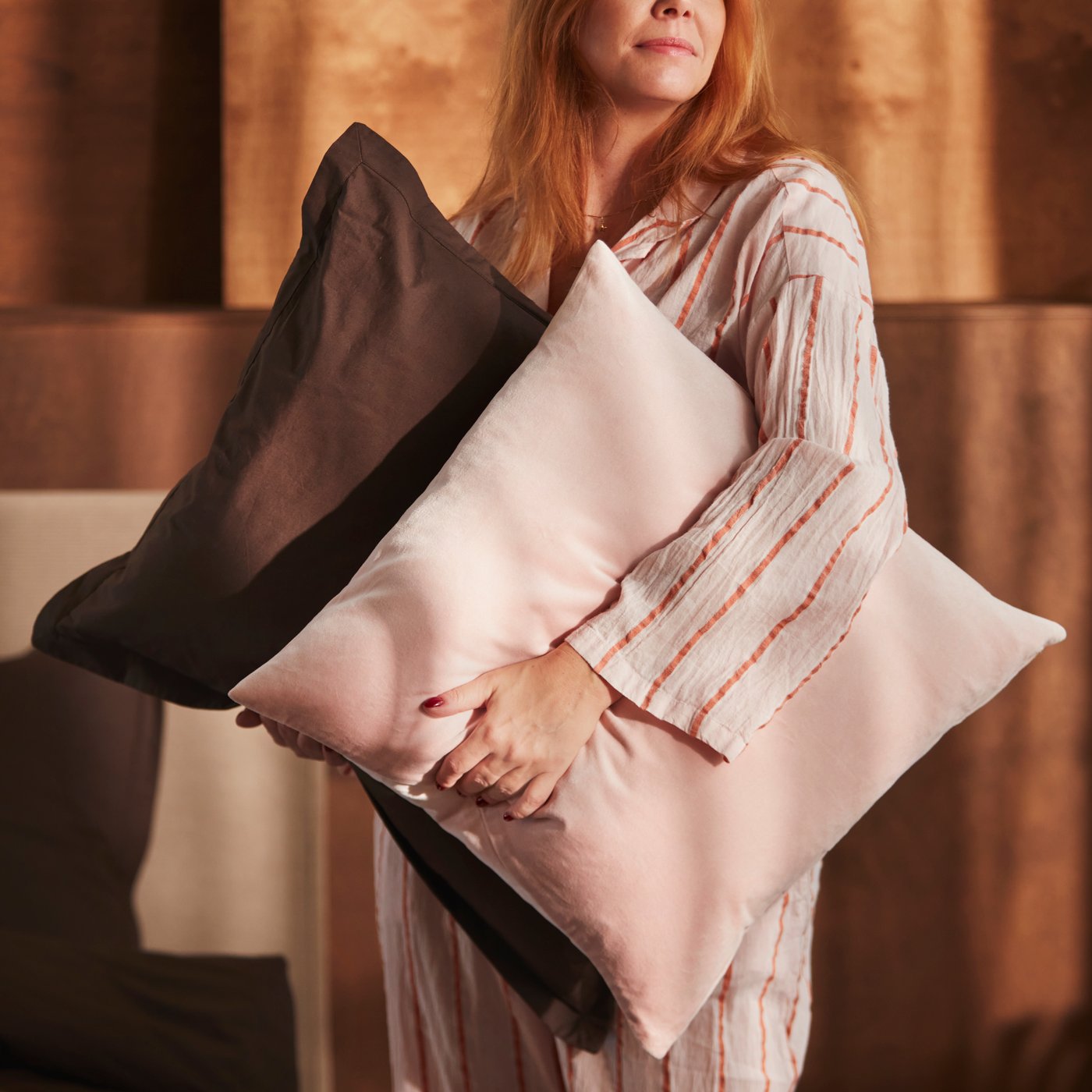 A person is holding a pillow with a dark brown HAMPFLOCKEL pillowcase and a cushion with a pale pink SANELA cushion cover.