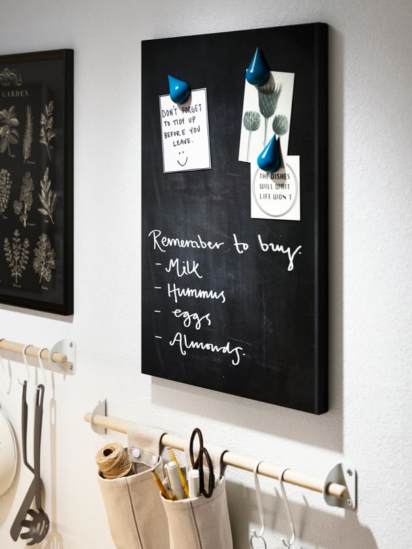 A wall section with a black SVENSÅS memo board with writing and notes. Below it hang accessories on a wall-mounted rail.