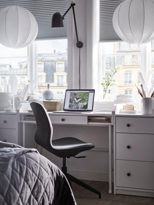 A white HAUGA desk between two chest of drawer units by a window with an open laptop on top and a work chair in front.