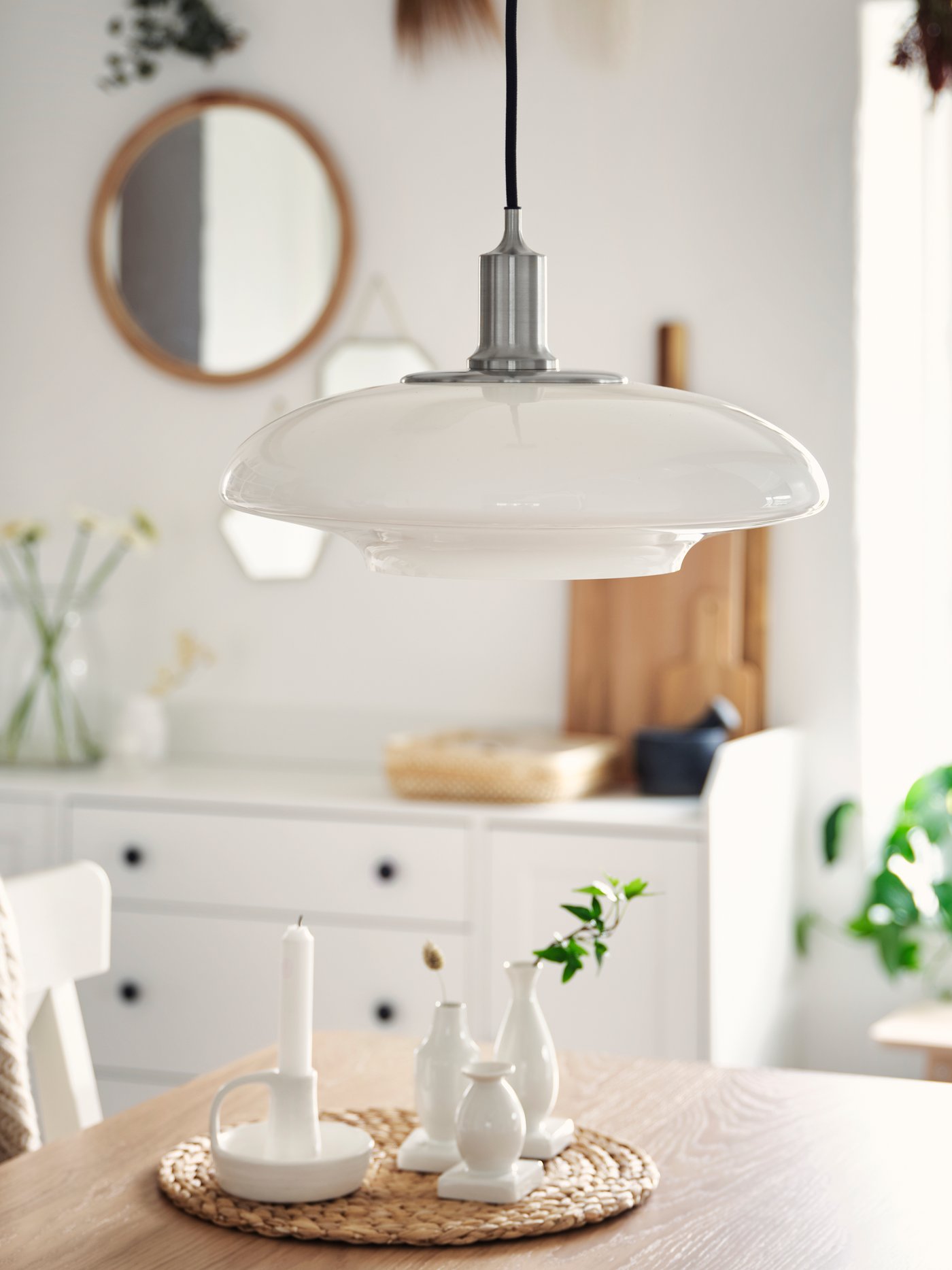 A nickel-plated pendant lamp with a white lampshade in glass hangs over a tabletop in oak veneer with white vases.