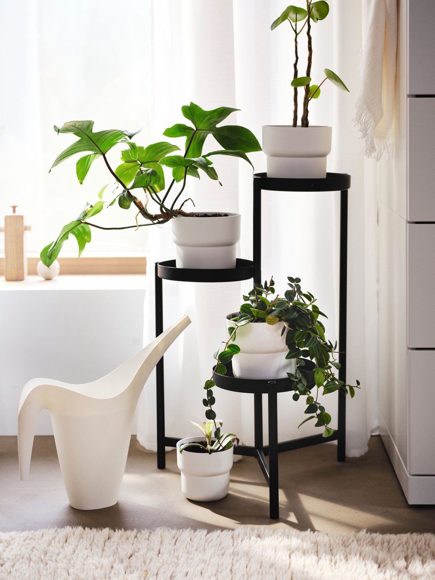 Regadera IKEA PS 2002 blanca junto a una base para plantas OLIVBLAD negra con macetas FÖRENLIG blancas y plantas, en un espacio interior.