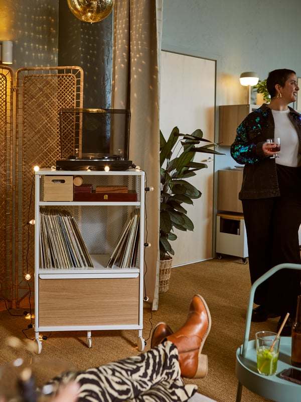 Espace à l’éclairage doux avec des personnes détendues se tenant près d’un rangement BEKANT blanc accueillant des disques vinyle et un tourne-disque.