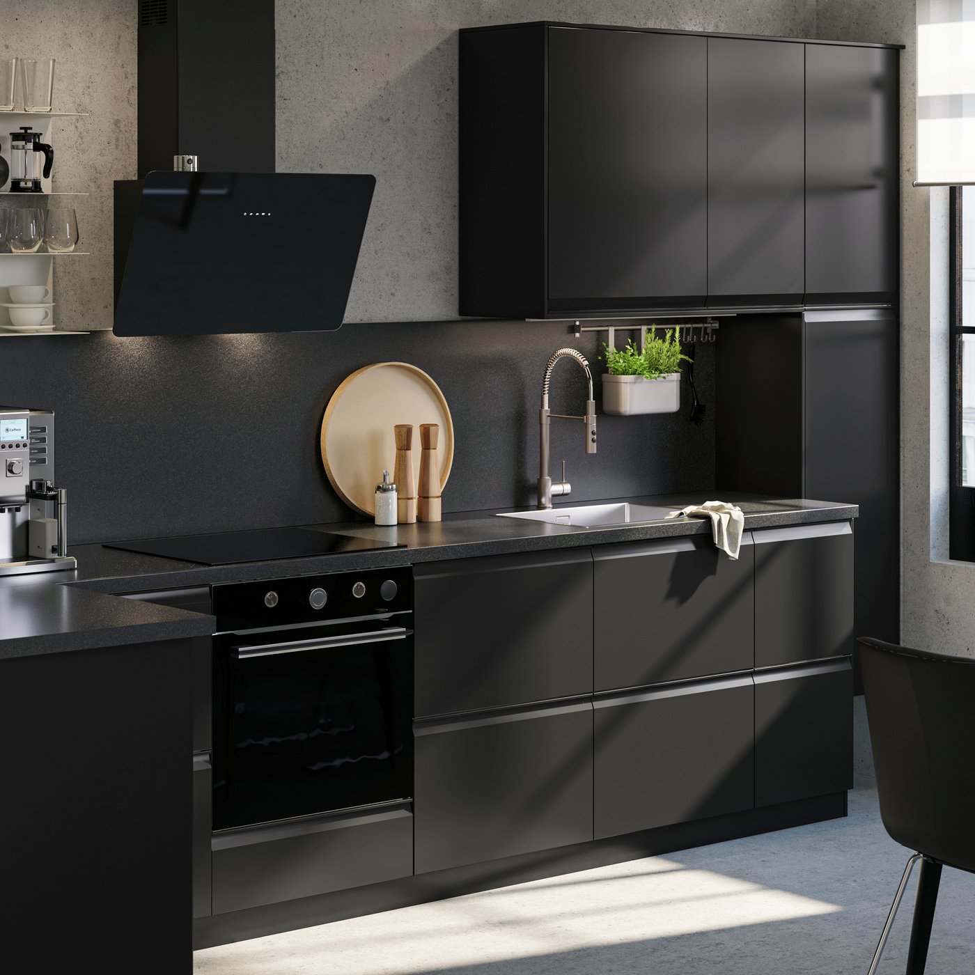 Moderní kuchyň IKEA s černými matnými dvířky, vestavnou troubou a varnou deskou, černým odsavačem par, nerezovým dřezem s baterií, závěsným květináčem, kávovarem na pracovní desce a jídelním stolem se čtyřmi židlemi. Nad stolem visí přírodní závěsné svítidlo IKEA SINNERLIG.
