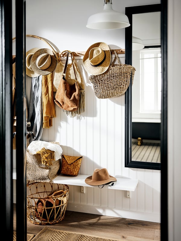 Un hall d’entrée blanc, des patères à 6 boutons où sont accrochés des chapeaux, sacs et foulards dans des coloris naturels. Des paniers sur une tablette et sur le sol.