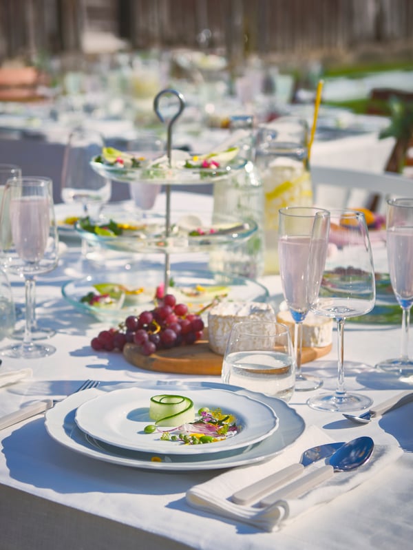 Ein Tisch ist mit GULLMAJ Tischdecke, DYRGRIP Gläsern, UPPLAGA Tellern und UPPHÖJD Besteck für eine Party im Freien gedeckt.