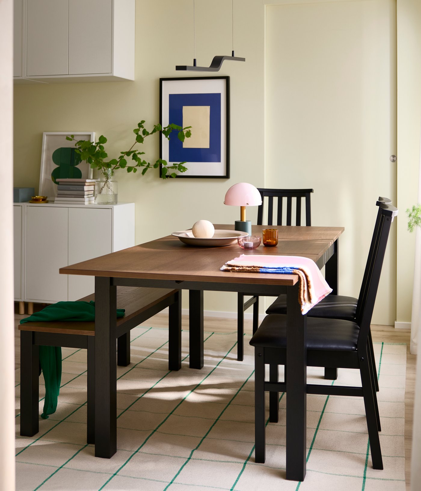 A bright dining area has an ÅLHULT extendable table in black/brown, with a matching bench and three chairs around it.