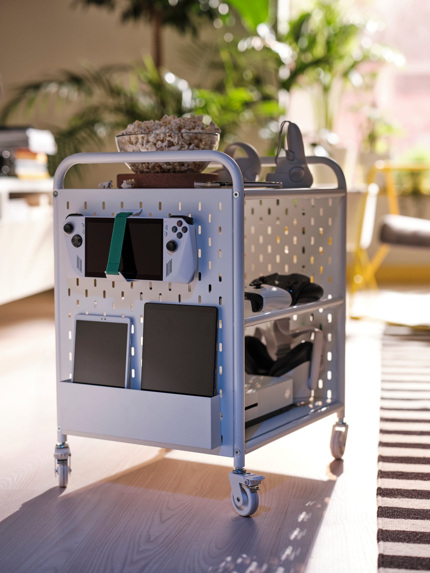 Estante BRÄNNBOLL blanco con ruedas, con un tazón de palomitas y accesorios gamer, además de dispositivos organizados en el panel perforado.