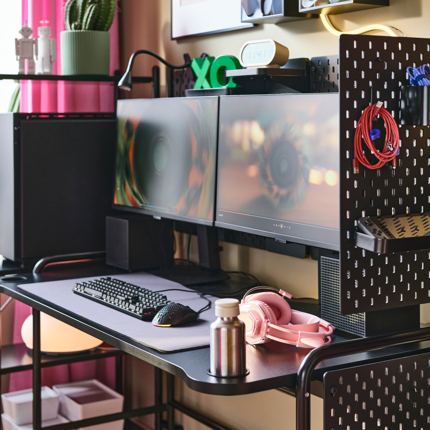 A dimly sunlit deskspace with a two-monitor setup centred around a black FRÖSKELÅS gaming desk lined with SKÅDIS pegboards.
