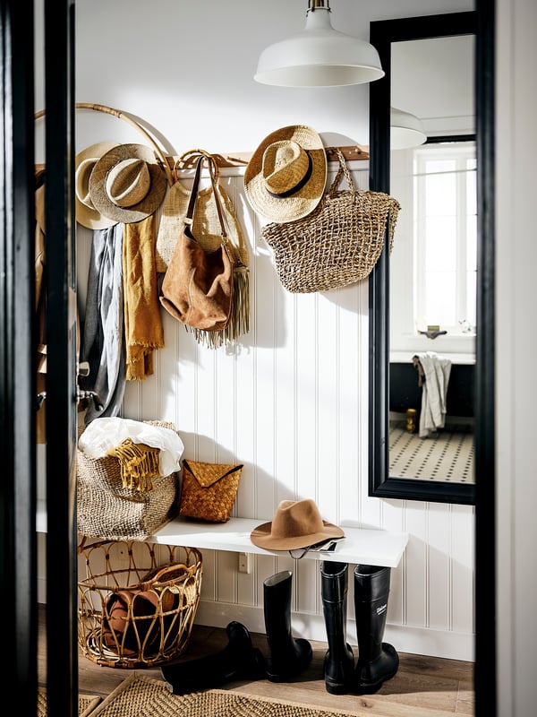 Un pasillo con bolsos, sombreros y bufandas de colores naturales en soportes con seis pomos y en canastas, junto a un espejo TOFTBYN negro