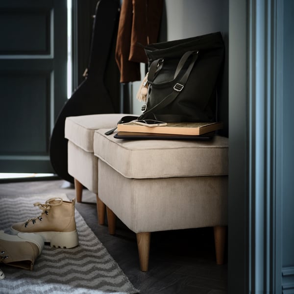 Two beige STRANDMON footstools line a hallway. Beneath them lie a pair of boots on a beige zigzag-patterned BREDEVAD rug.