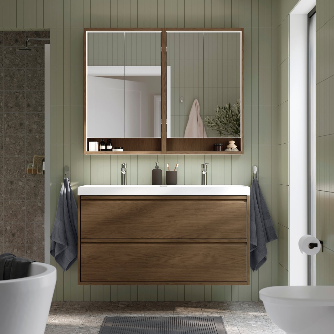 Warm bathroom with a wood vanity, dual faucets and mirrored cabinets against green tiled walls.