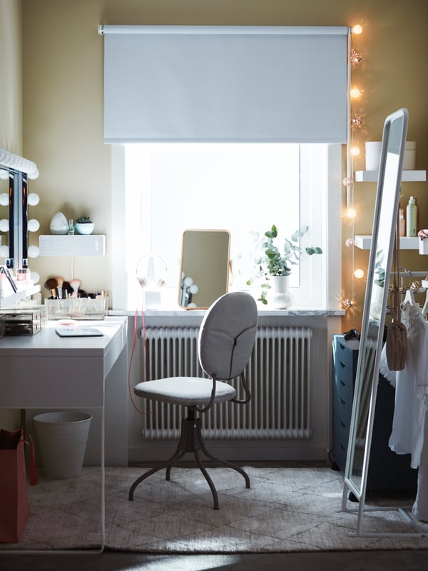 Une chaise pivotante beige avec un bureau MICKE blanc, à côté d’un miroir de table et une plante sur le rebord d’une fenêtre, avec un miroir sur pied blanc.