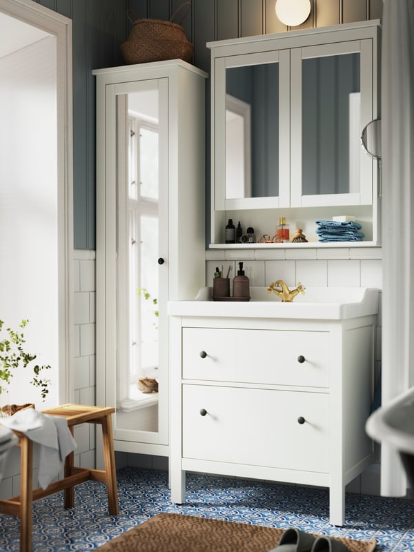 A white HEMNES vanity with two drawers and a high cabinet with a mirror door are standing in a blue and white bathroom.