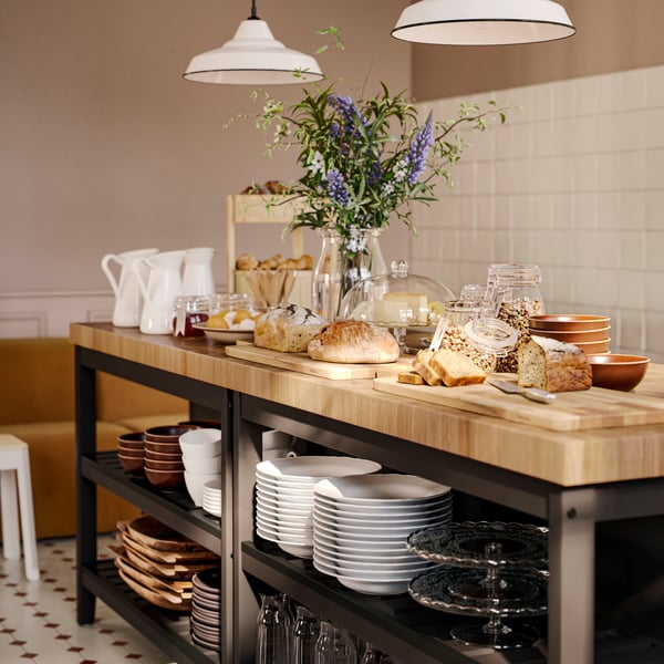 Un buffet della colazione con vari prodotti, tra cui dei contenitori con coperchio KORKEN contenenti confettura e granola, è servito su due isole per cucina in legno.