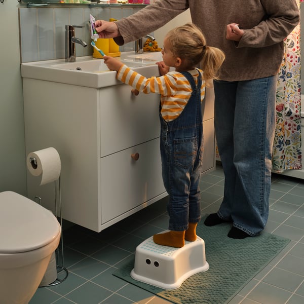 A parent helps a toddler brushing the teeth, the toddler standing on FÖRSIKTIG children’s stool to reach up to the sink.