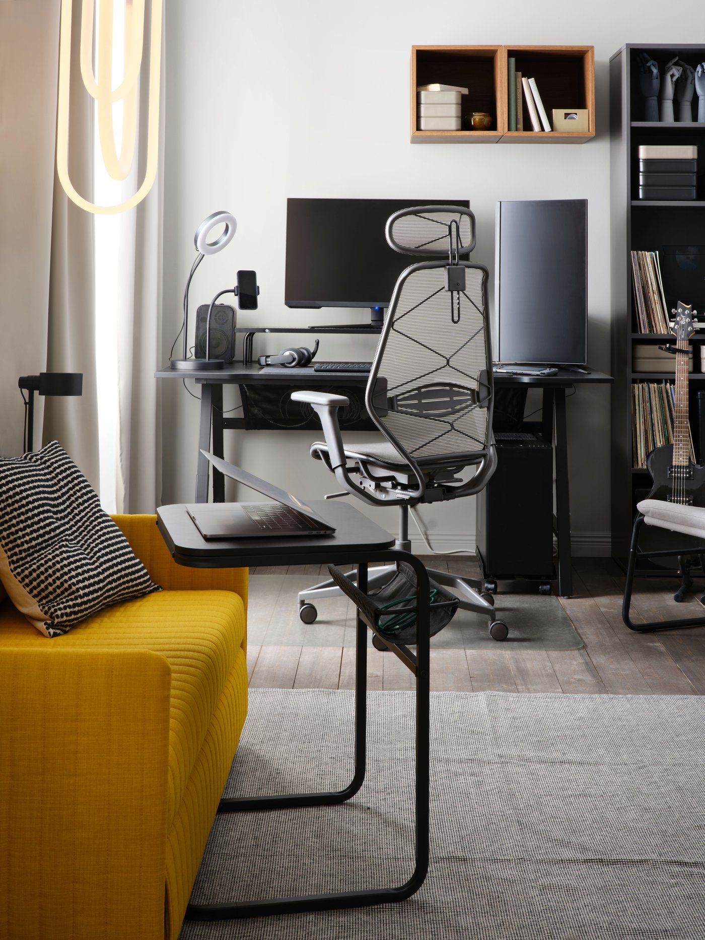 A dark grey/grey STYRSPEL gaming chair is by a black gaming desk. The foreground has a yellow sofa and a dark grey side table.